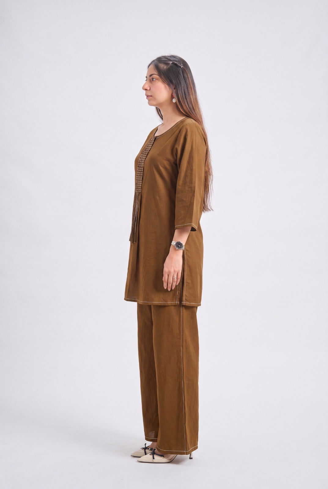 A woman stands in profile wearing a brown two-piece kurta and trouser set against a plain light gray studio background.