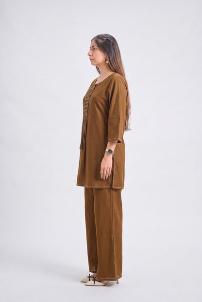 A woman stands in profile wearing a brown two-piece kurta and trouser set against a plain light gray studio background.