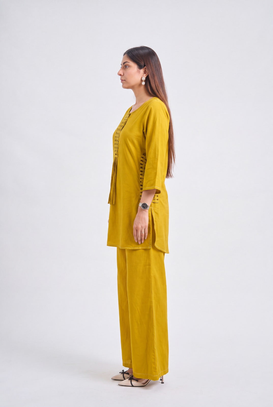 A woman stands in profile wearing a mustard yellow two-piece outfit with a short-sleeve top and wide-leg pants, photographed against a plain light gray studio background.