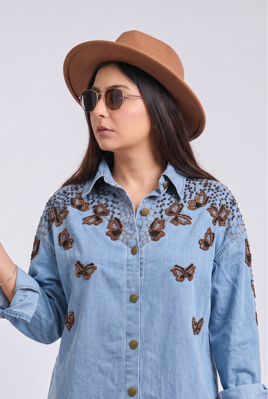 A woman wearing a light blue denim button-up shirt with brown butterfly embroidery on the chest and shoulders, paired with a brown hat, sunglasses, and jeans, holding a coffee cup against a light gray background.