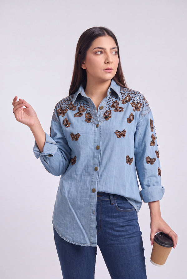 A woman modeling a light blue denim button-up shirt with brown butterfly embroidery on the shoulders and sleeves, paired with dark blue jeans, holding a coffee cup against a light gray background.