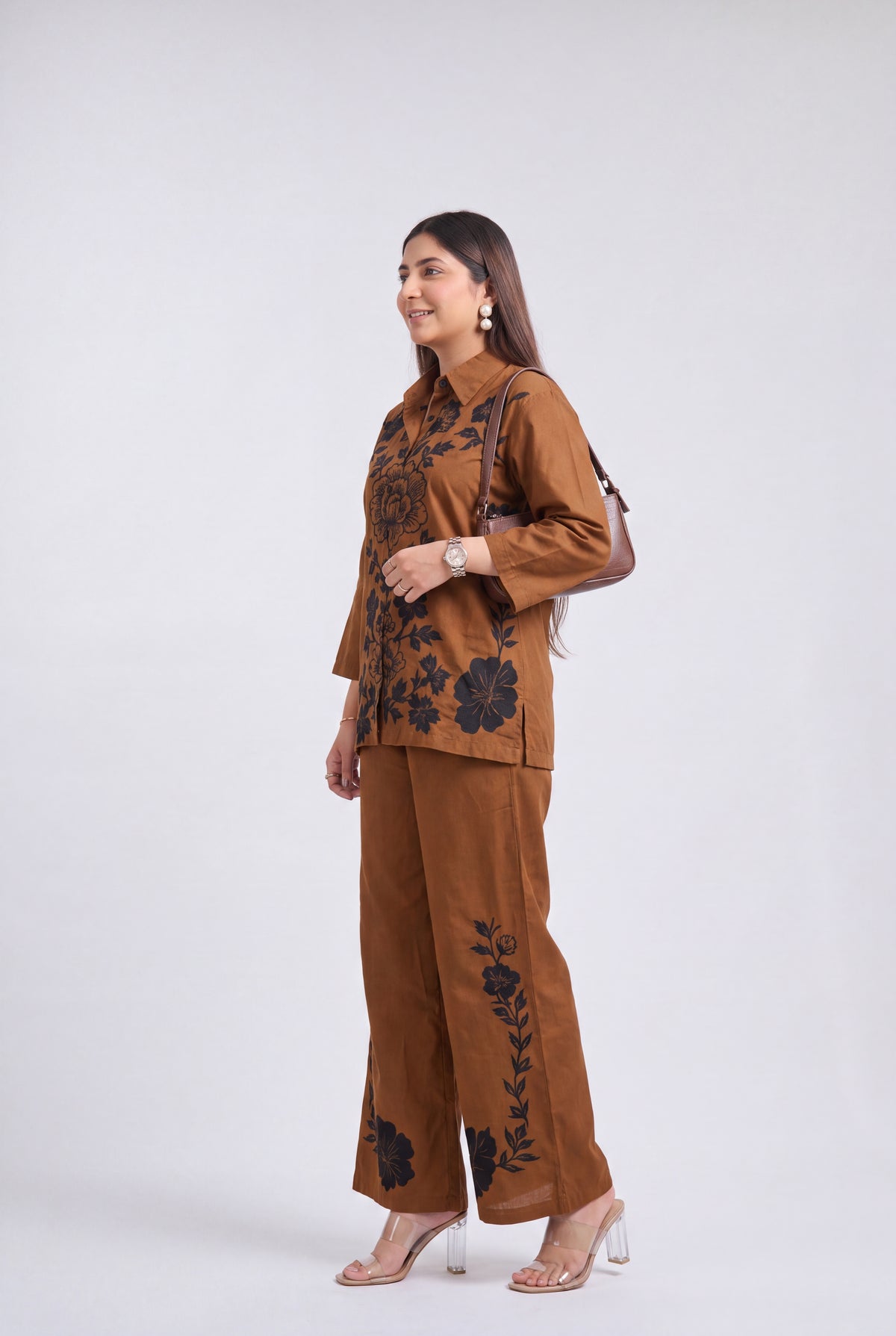 A woman modeling a brown two-piece set consisting of a long-sleeve button-up top and matching wide-leg trousers, both decorated with black floral embroidery, set against a plain light gray background.