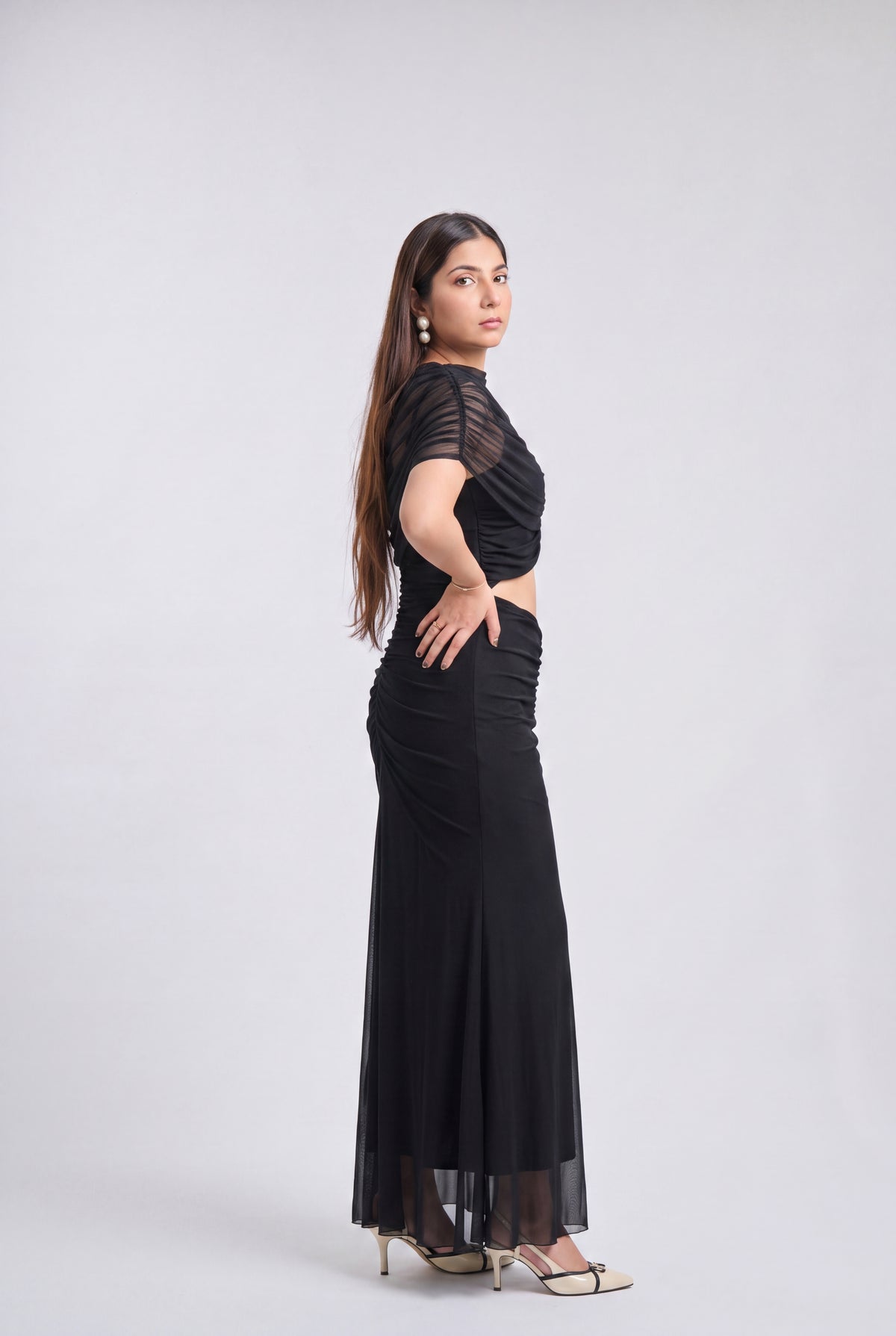 A woman modeling a black floor-length evening gown with sheer short sleeves and a draped, ruched bodice, shown in profile against a light gray studio background.