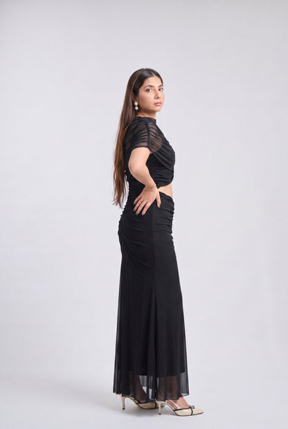 A woman modeling a black floor-length evening gown with sheer short sleeves and a draped, ruched bodice, shown in profile against a light gray studio background.