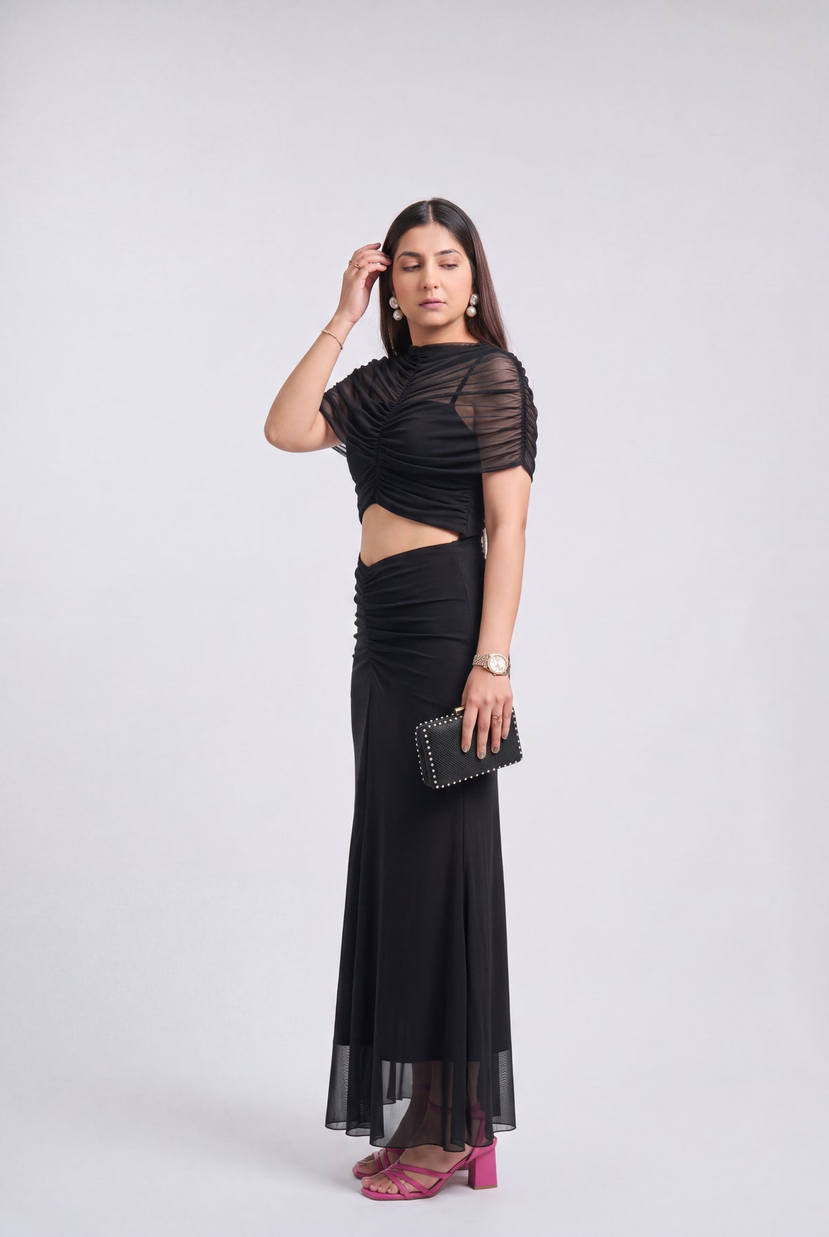 A woman modeling a black two-piece outfit consisting of a short-sleeve cropped top with sheer mesh sleeves and a matching long ruched skirt, paired with pink heels and a black clutch, against a light gray studio background.