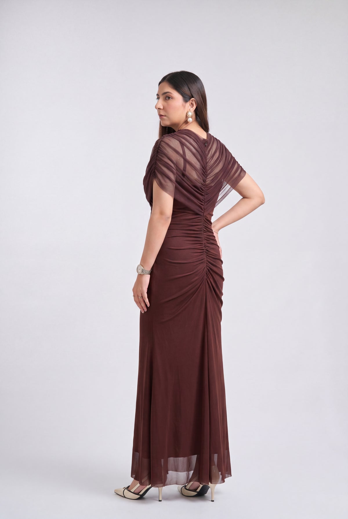 A woman modeling a floor-length, dark brown ruched gown with sheer short sleeves and draped detailing, shown from the back against a light gray studio background.