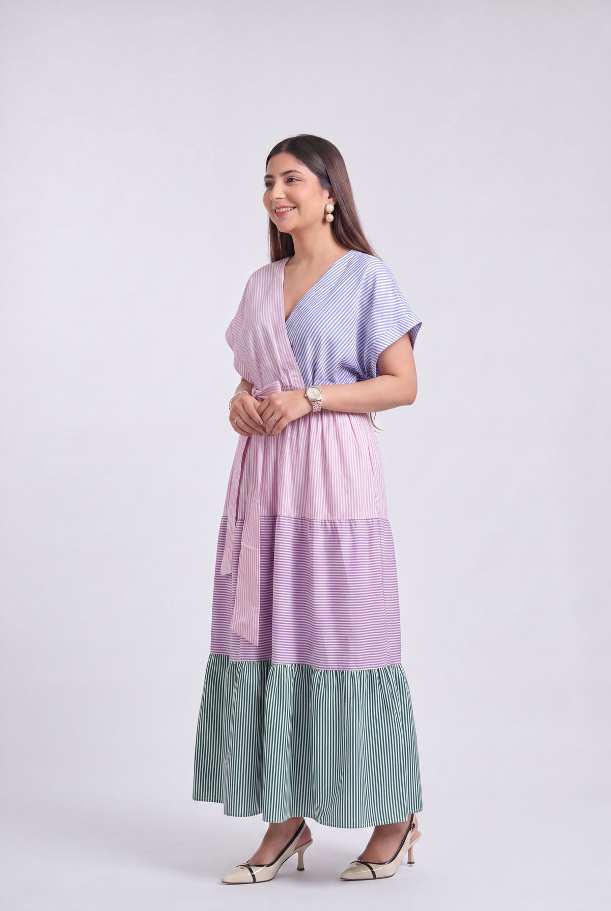 A woman modeling a long, tiered wrap dress with a lavender and light blue striped pattern and a sage green pleated ruffled hem, set against a plain light gray studio background.