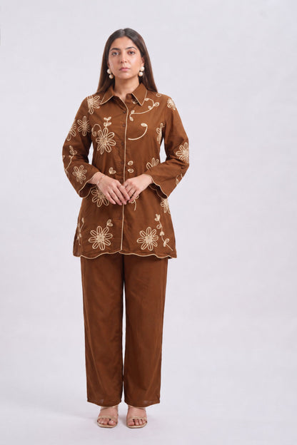 A woman modeling a two-piece pajama set consisting of a brown long-sleeve button-up shirt and matching wide-leg pants, both decorated with beige floral embroidery, set against a plain light gray background.