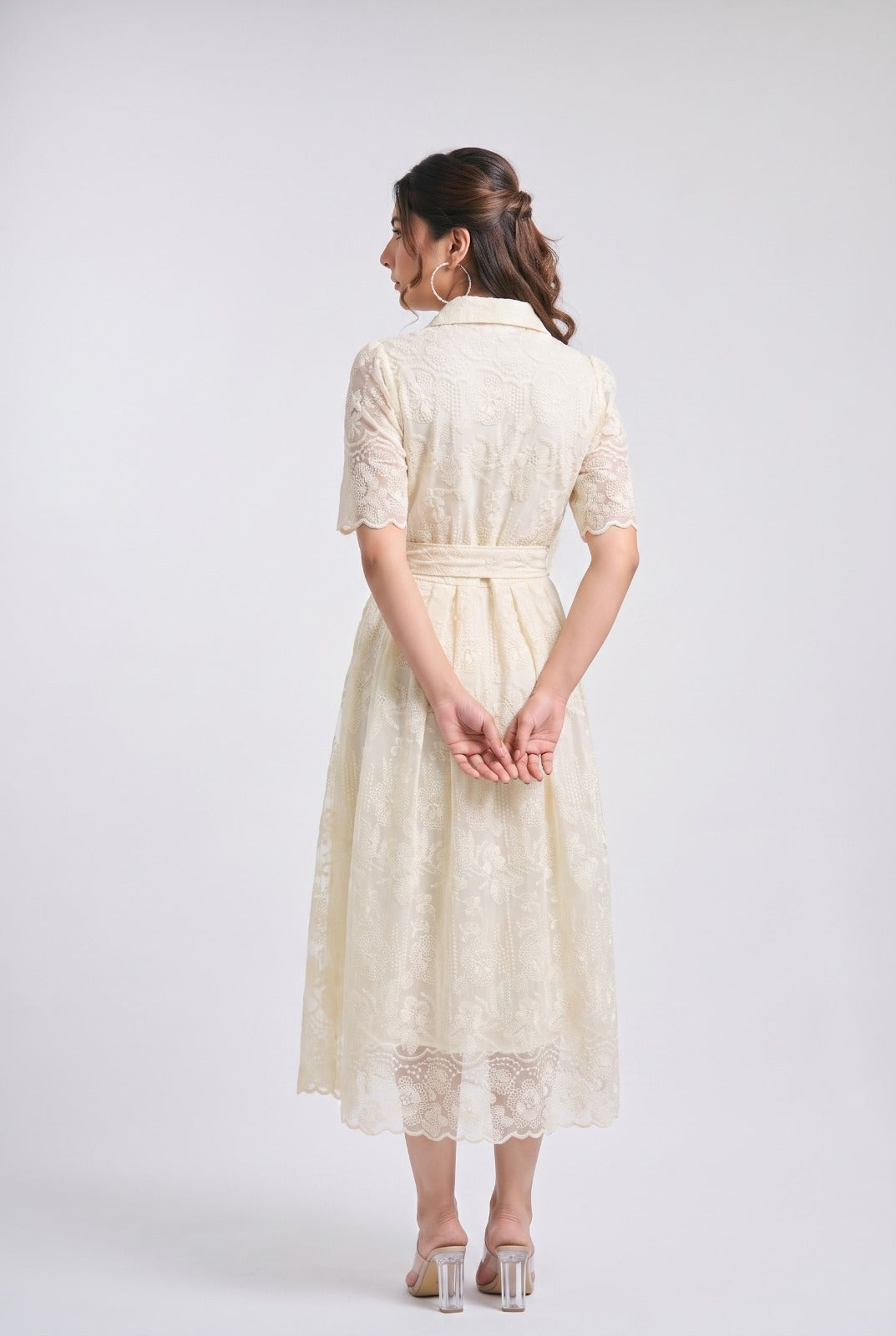 A woman is shown from the back wearing a cream-colored lace midi dress with short sleeves and a matching waist tie, paired with clear high-heeled sandals, against a light gray studio background.