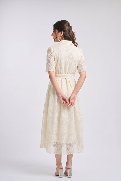 A woman is shown from the back wearing a cream-colored lace midi dress with short sleeves and a matching waist tie, paired with clear high-heeled sandals, against a light gray studio background.