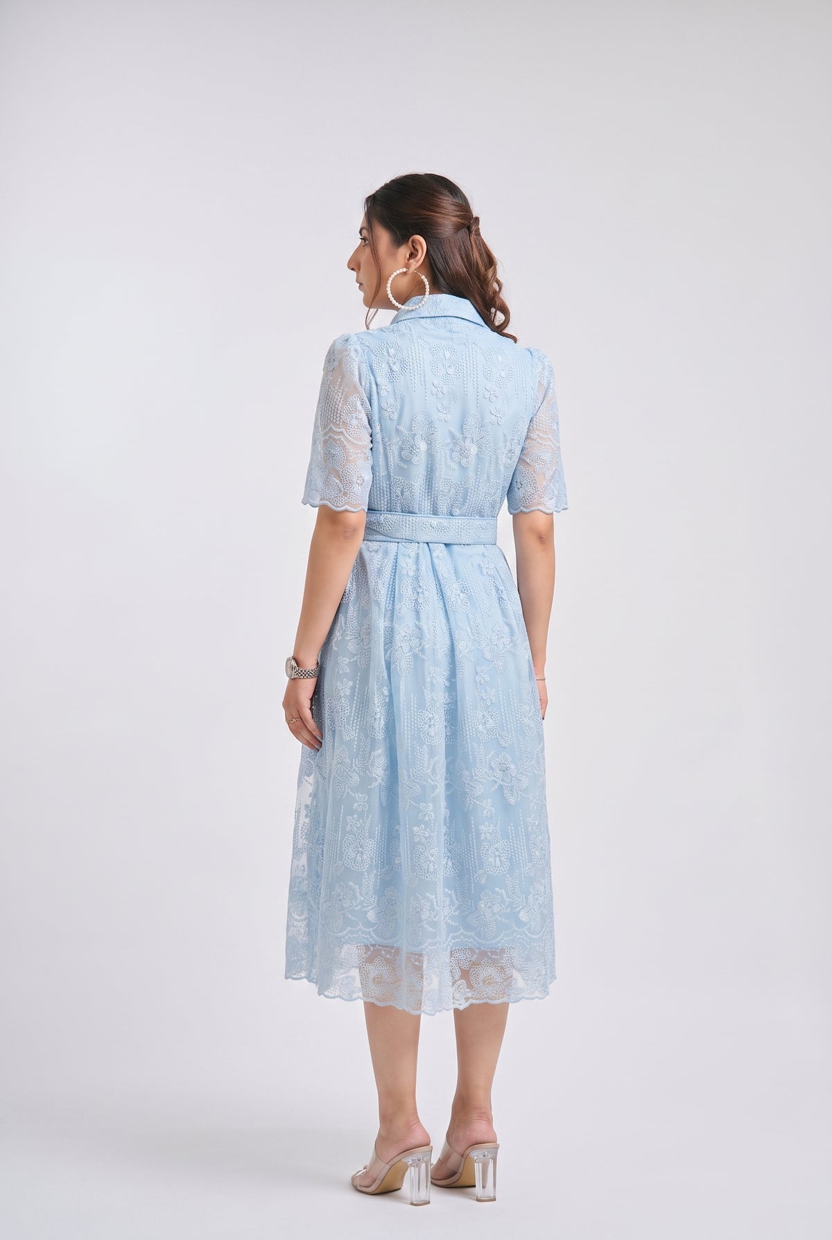 A woman viewed from the back wearing a light blue lace midi dress with short sleeves, a collar, and a matching waist tie, paired with clear high-heeled sandals against a light gray studio background.