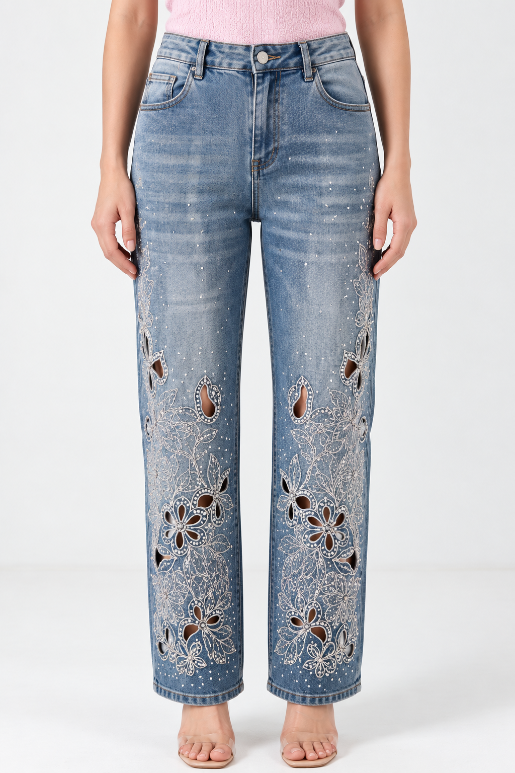 A model wearing light-wash blue jeans with intricate white floral embroidery and cutout details on the front of the legs, paired with a pink top against a white studio background.