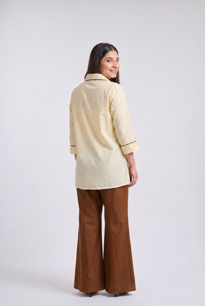 A woman is shown from the back wearing a cream-colored cotton kurta with dark piping and brown wide-leg pants, standing against a light gray studio background.