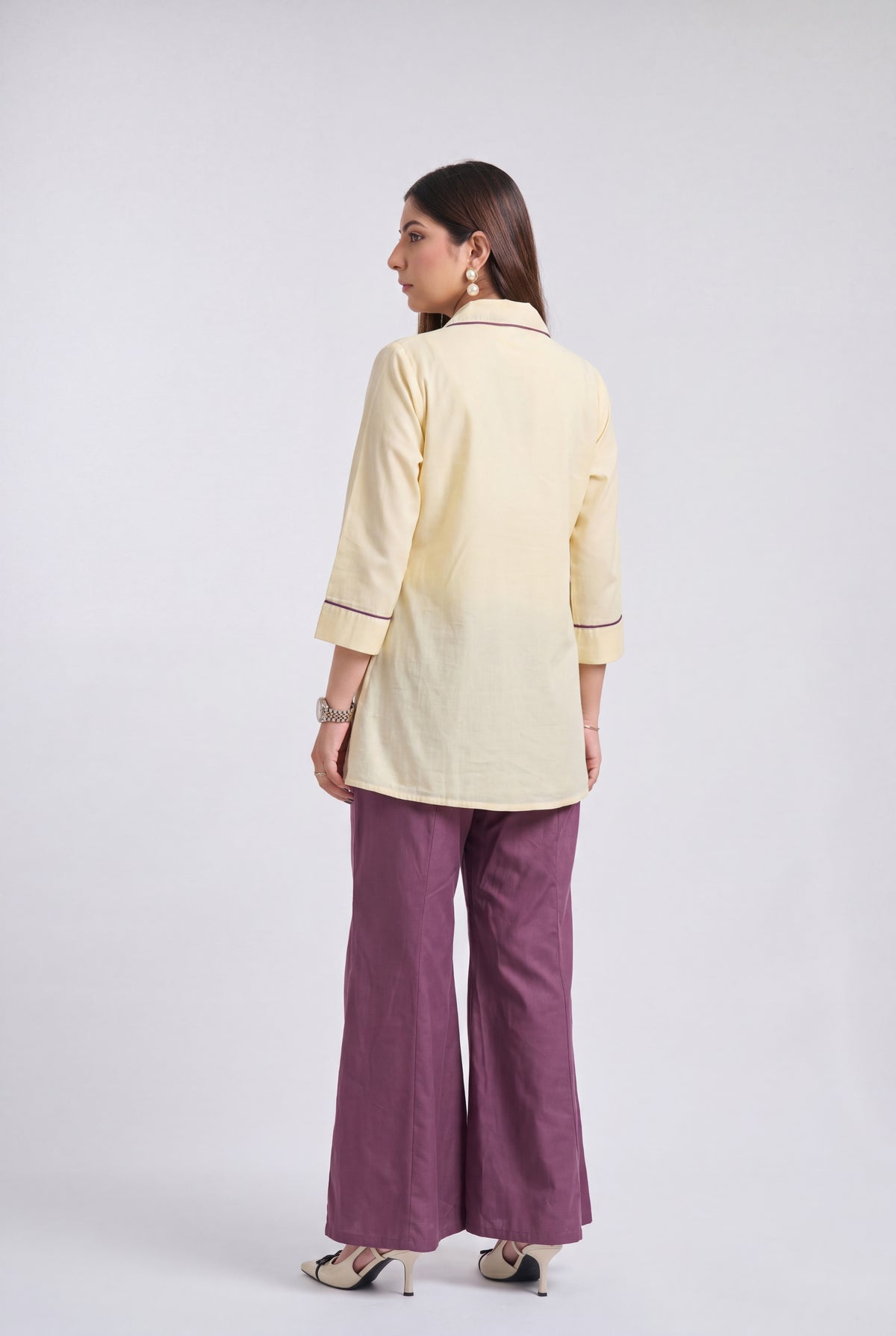 A woman is shown from the back wearing a pale yellow three-quarter sleeve kurta with thin dark piping details, paired with wide-leg purple trousers and beige heels, against a light gray studio background.