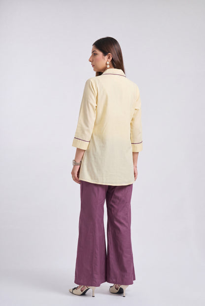 A woman is shown from the back wearing a pale yellow three-quarter sleeve kurta with thin dark piping details, paired with wide-leg purple trousers and beige heels, against a light gray studio background.