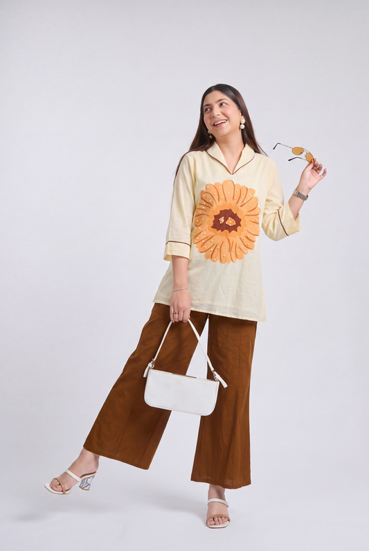 A woman modeling a cream-colored long-sleeve top with a large orange sunflower graphic on the front, paired with brown wide-leg pants, a white handbag, and white heeled sandals, set against a plain light gray studio background.
