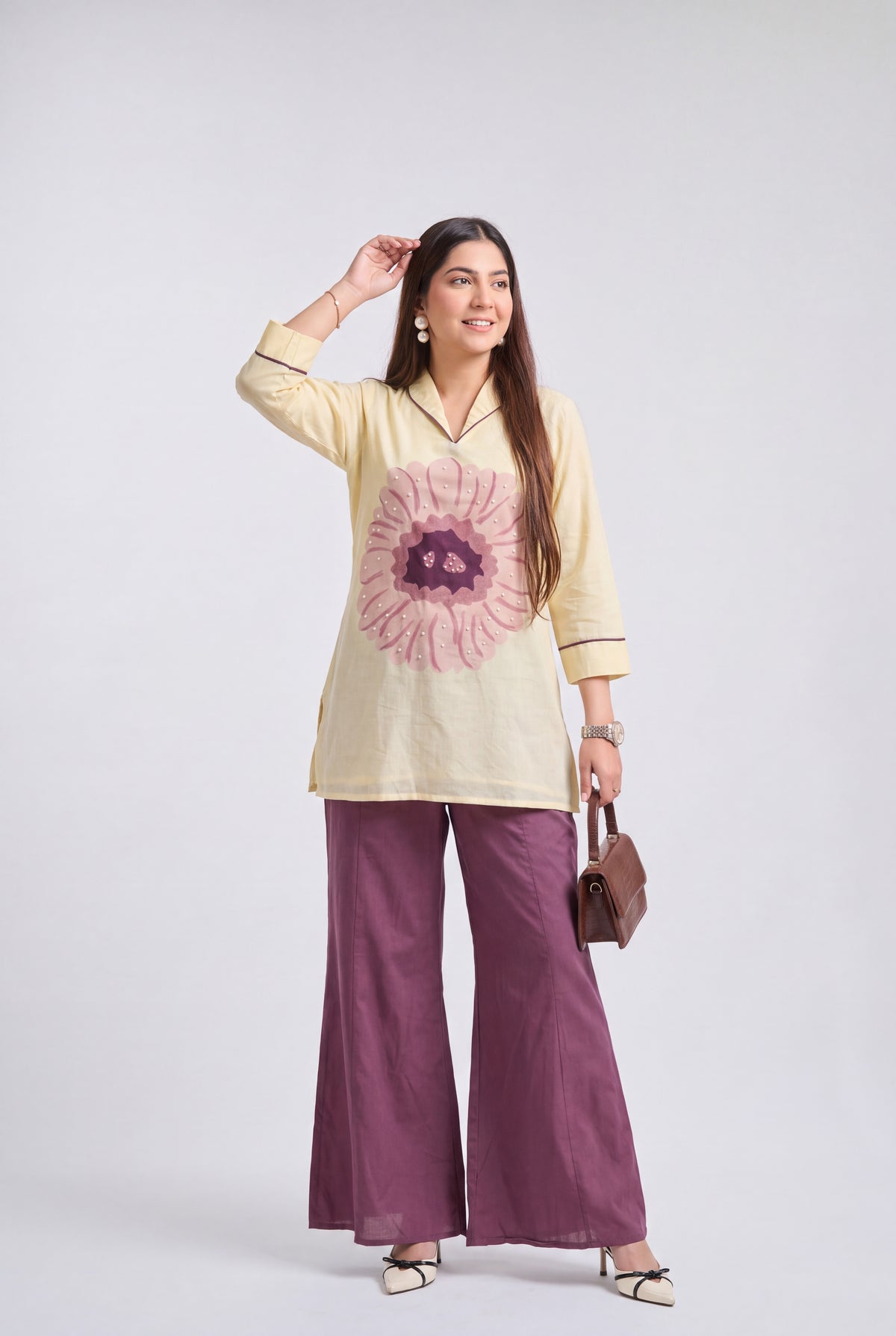 A woman modeling a cream-colored tunic top with a pink and purple floral print, paired with wide-leg purple trousers, holding a small brown handbag against a light gray studio background.