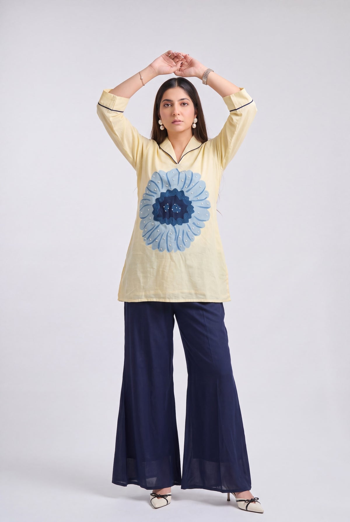 A woman modeling a cream-colored kurta with a large blue floral print on the front, paired with navy blue wide-leg pants, against a light gray studio background.
