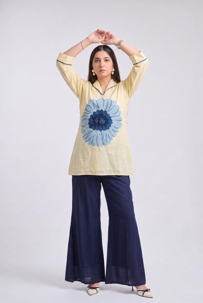 A woman modeling a cream-colored kurta with a large blue floral print on the front, paired with navy blue wide-leg pants, against a light gray studio background.
