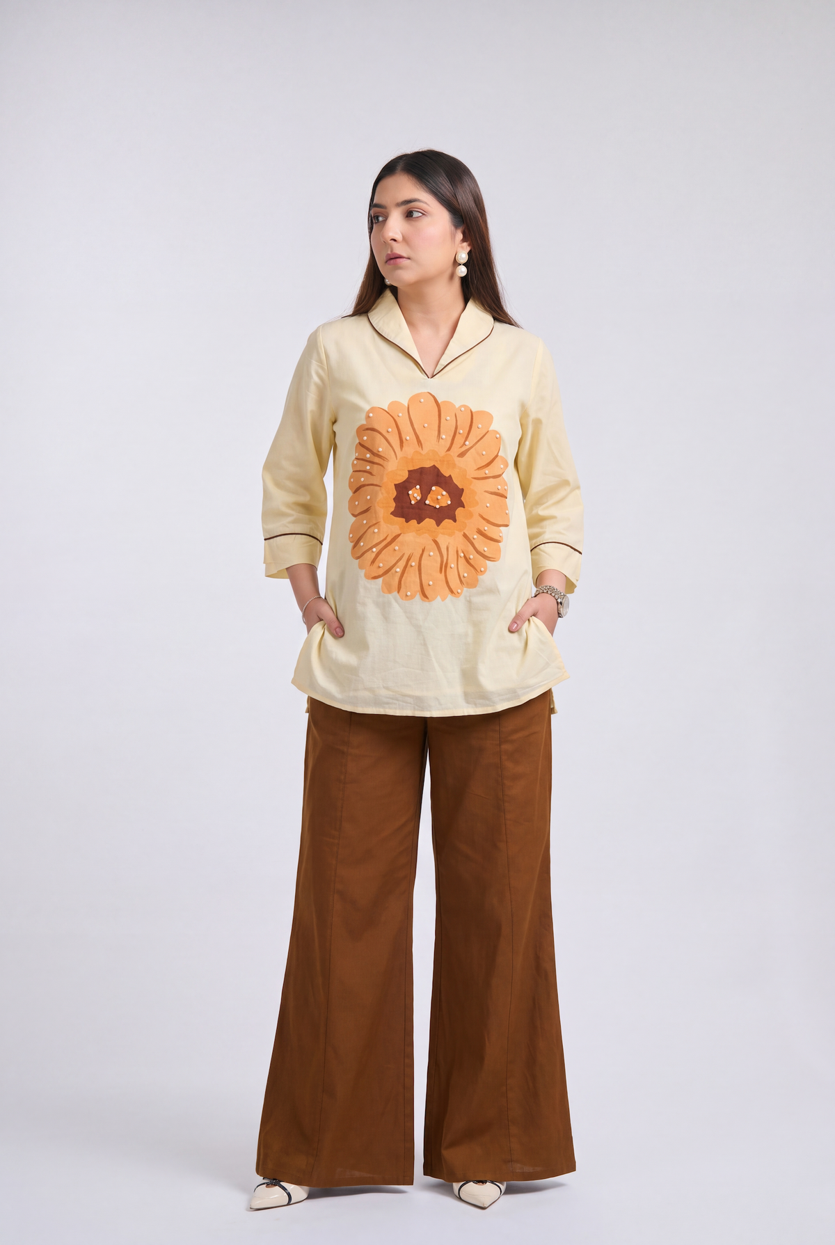 A woman modeling a cream-colored long-sleeve shirt with a large orange sunflower graphic on the front, paired with brown wide-leg pants, against a light gray studio background.