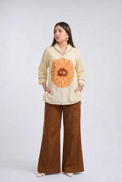 A woman modeling a cream-colored long-sleeve shirt with a large orange sunflower graphic on the front, paired with brown wide-leg pants, against a light gray studio background.