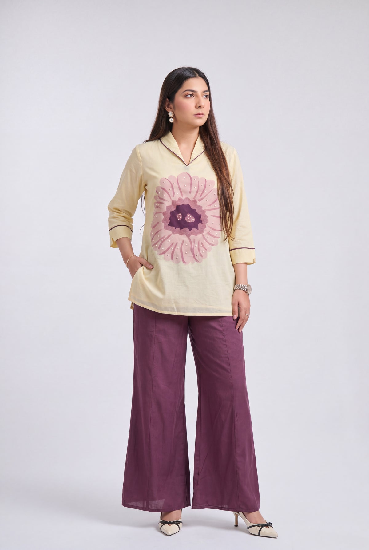 A woman modeling a cream-colored cotton kurta with a large pink floral print on the front, paired with wide-leg purple trousers and beige heels, set against a plain light gray studio background.