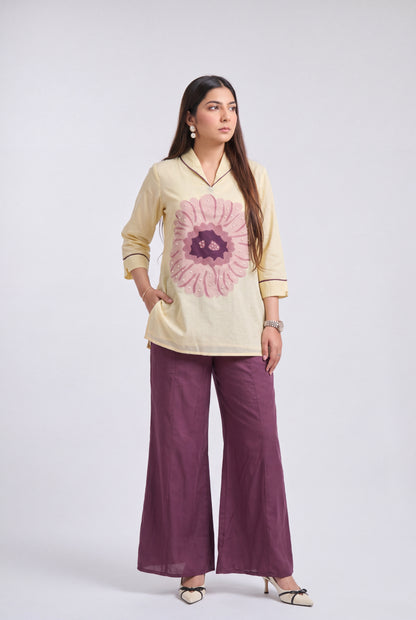 A woman modeling a cream-colored cotton kurta with a large pink floral print on the front, paired with wide-leg purple trousers and beige heels, set against a plain light gray studio background.