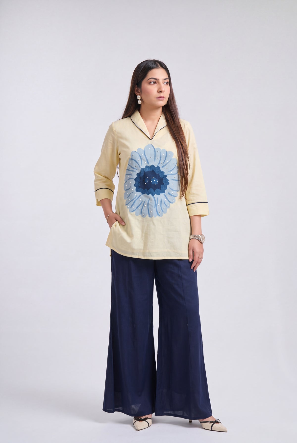 A woman modeling a light beige long-sleeve kurta with a large blue floral print on the front, paired with navy blue wide-leg trousers, against a light gray studio background.