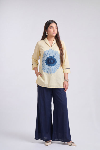 A woman modeling a light beige long-sleeve kurta with a large blue floral print on the front, paired with navy blue wide-leg trousers, against a light gray studio background.