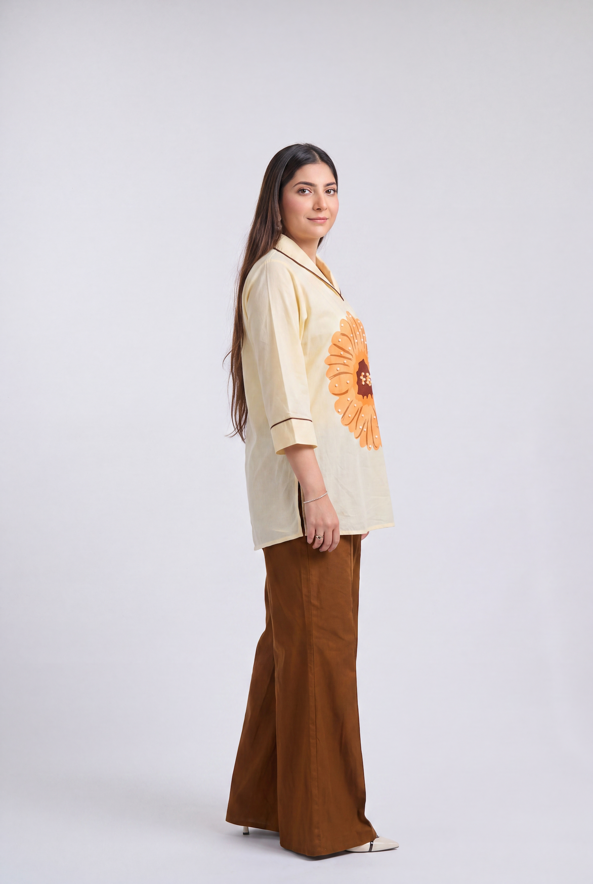 A woman stands in profile wearing a cream-colored short-sleeve top with a large orange sunflower graphic on the front and brown wide-leg pants, photographed against a light gray studio background.