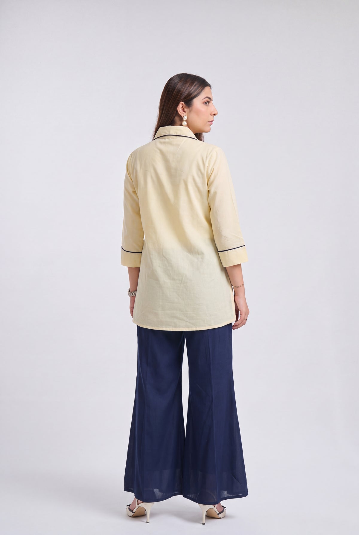 A woman is shown from the back wearing a light cream-colored kurta with dark piping details and navy blue wide-leg pants, standing against a light gray studio background.