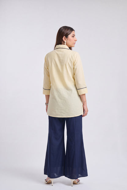 A woman is shown from the back wearing a light cream-colored kurta with dark piping details and navy blue wide-leg pants, standing against a light gray studio background.