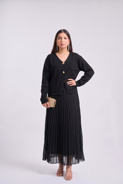 A woman modeling a black long-sleeve cardigan with gold buttons over a matching black pleated maxi skirt, holding a small gold clutch against a light gray studio background.