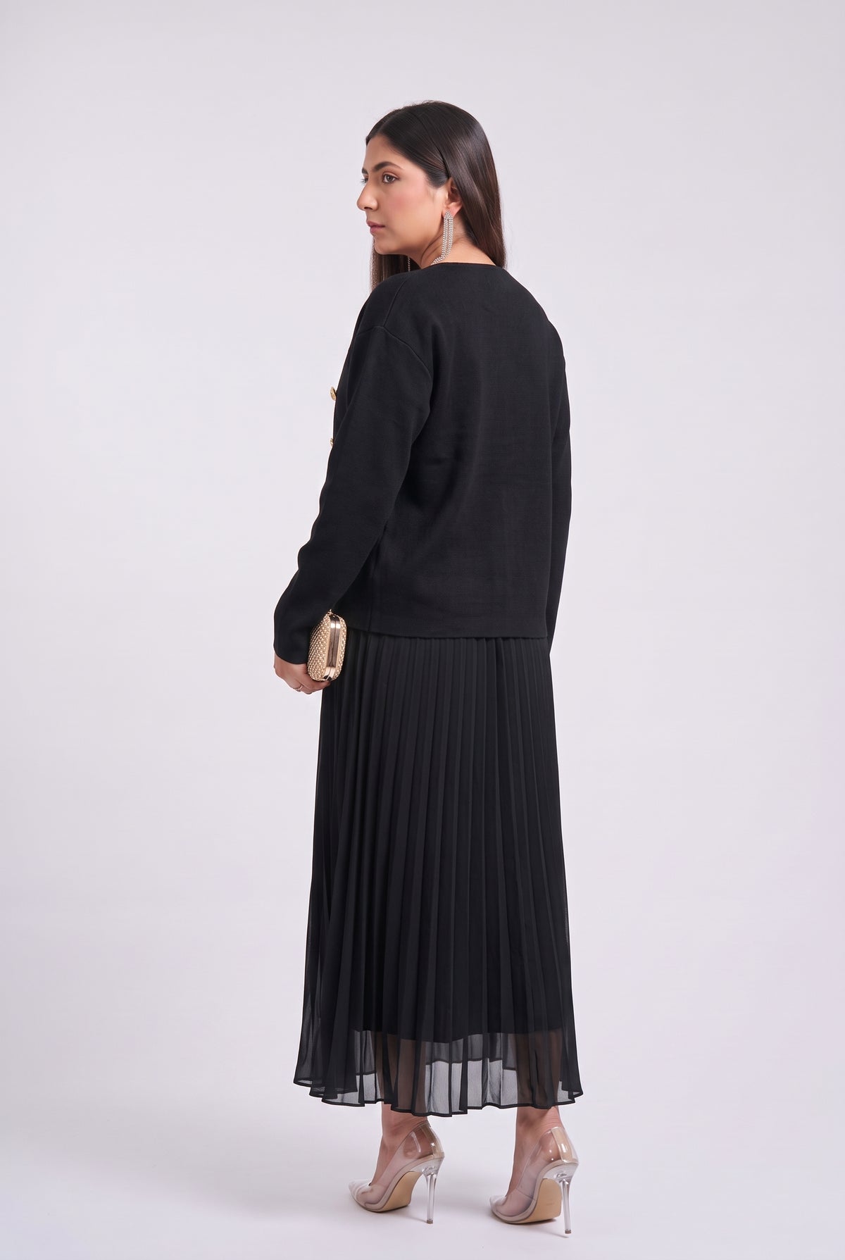 A woman is shown from the back wearing a black long-sleeve top and a black pleated midi skirt with a sheer hem, paired with clear high-heeled sandals and holding a small gold clutch, set against a plain light gray studio background.