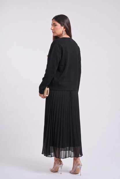 A woman is shown from the back wearing a black long-sleeve top and a black pleated midi skirt with a sheer hem, paired with clear high-heeled sandals and holding a small gold clutch, set against a plain light gray studio background.