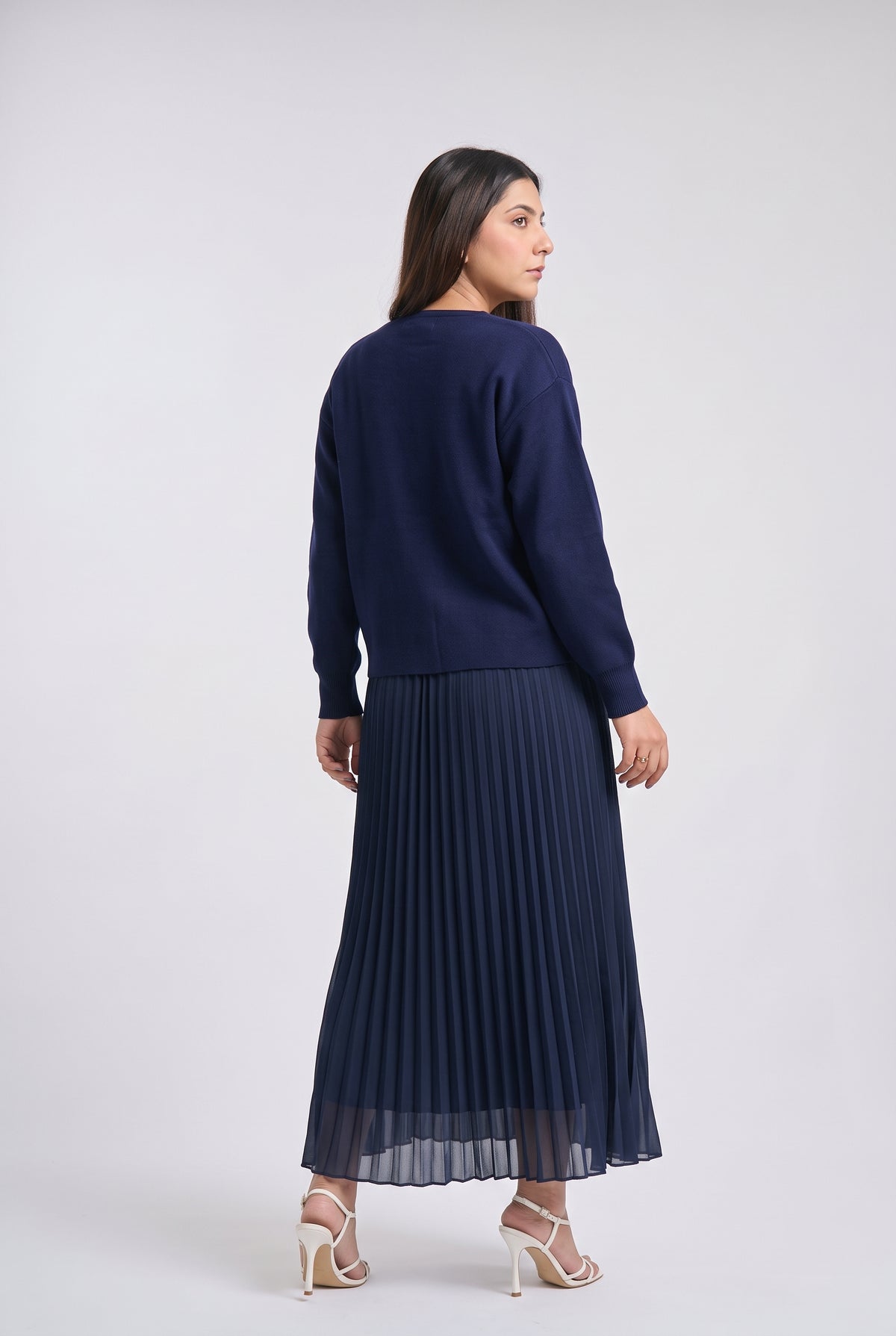 A woman is shown from the back wearing a navy blue knit sweater and a matching navy blue pleated maxi skirt, paired with white high-heeled sandals, against a light gray studio background.