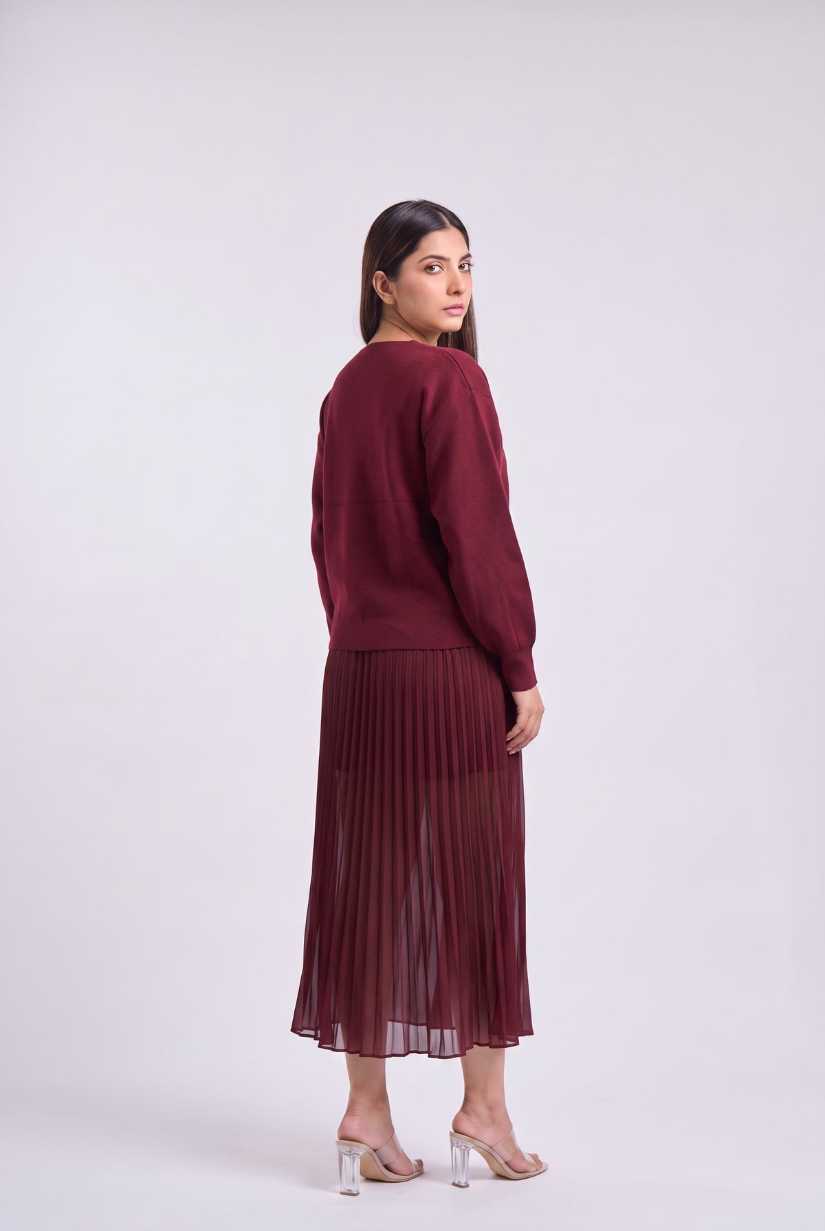A woman modeling a matching burgundy knit sweater and pleated midi skirt set, shown from a three-quarters back view against a light gray studio background.