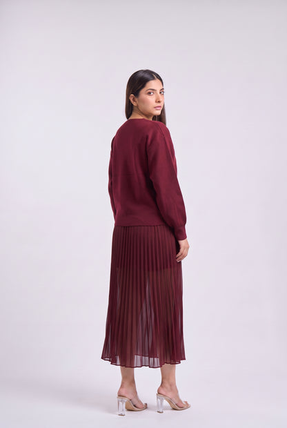A woman modeling a matching burgundy knit sweater and pleated midi skirt set, shown from a three-quarters back view against a light gray studio background.