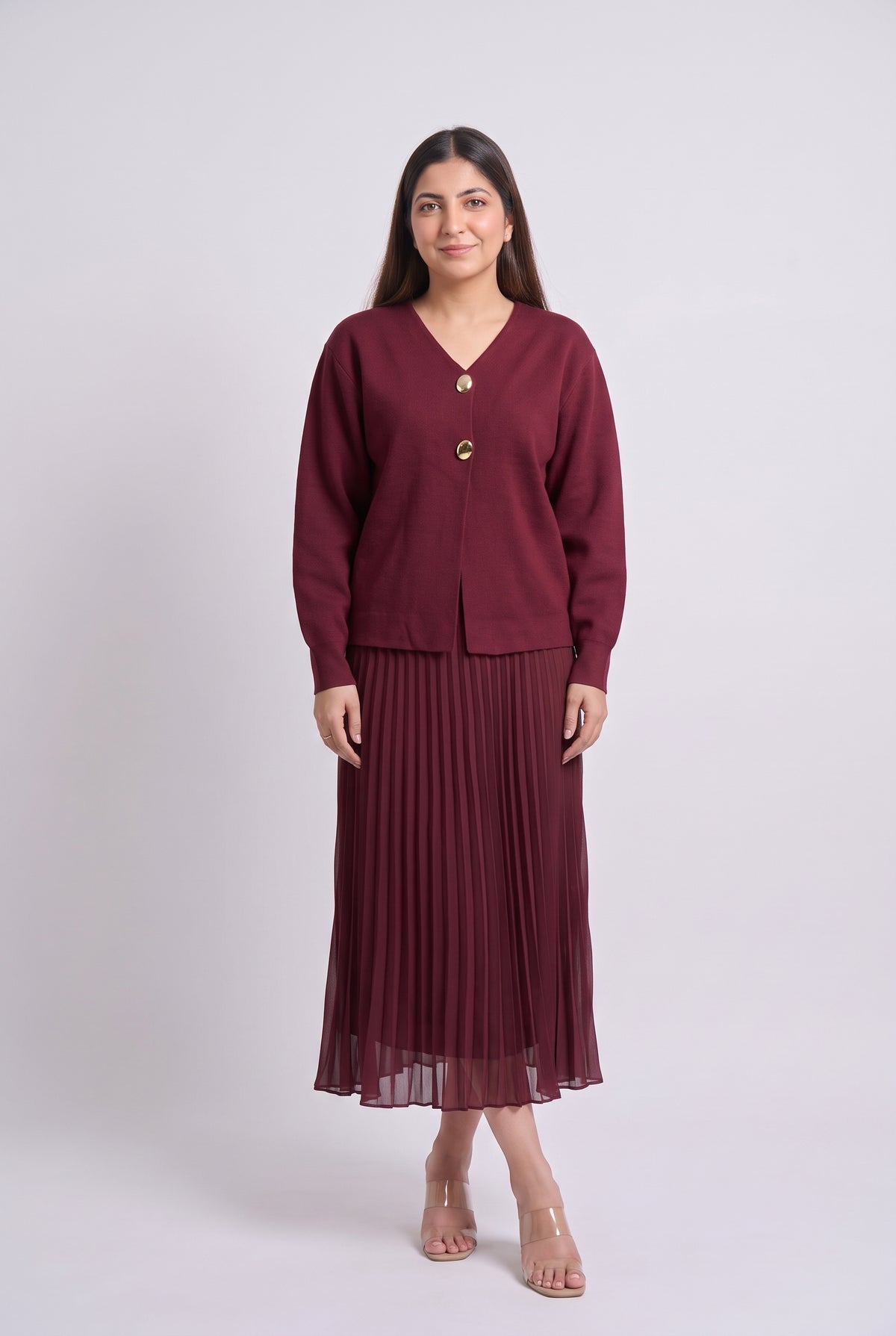 A woman modeling a matching burgundy knit cardigan and pleated midi skirt set with gold buttons, standing against a light gray studio background.