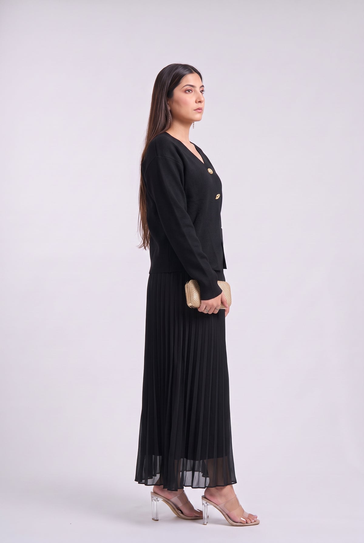A woman modeling a long black maxi dress with a double-breasted blazer-style bodice and pleated skirt, standing in profile against a light gray studio background.