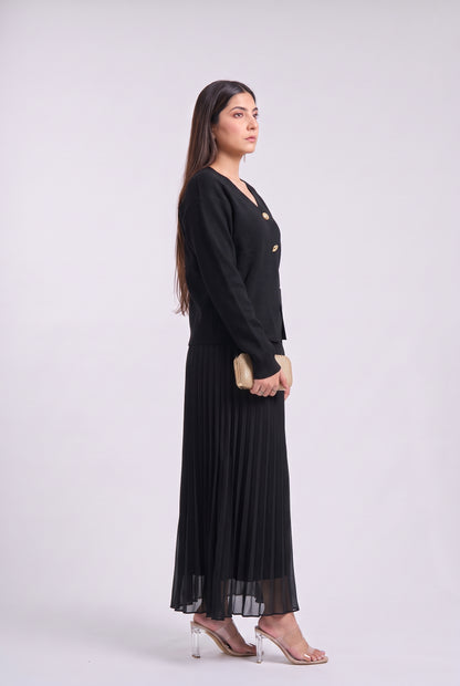 A woman modeling a long black maxi dress with a double-breasted blazer-style bodice and pleated skirt, standing in profile against a light gray studio background.
