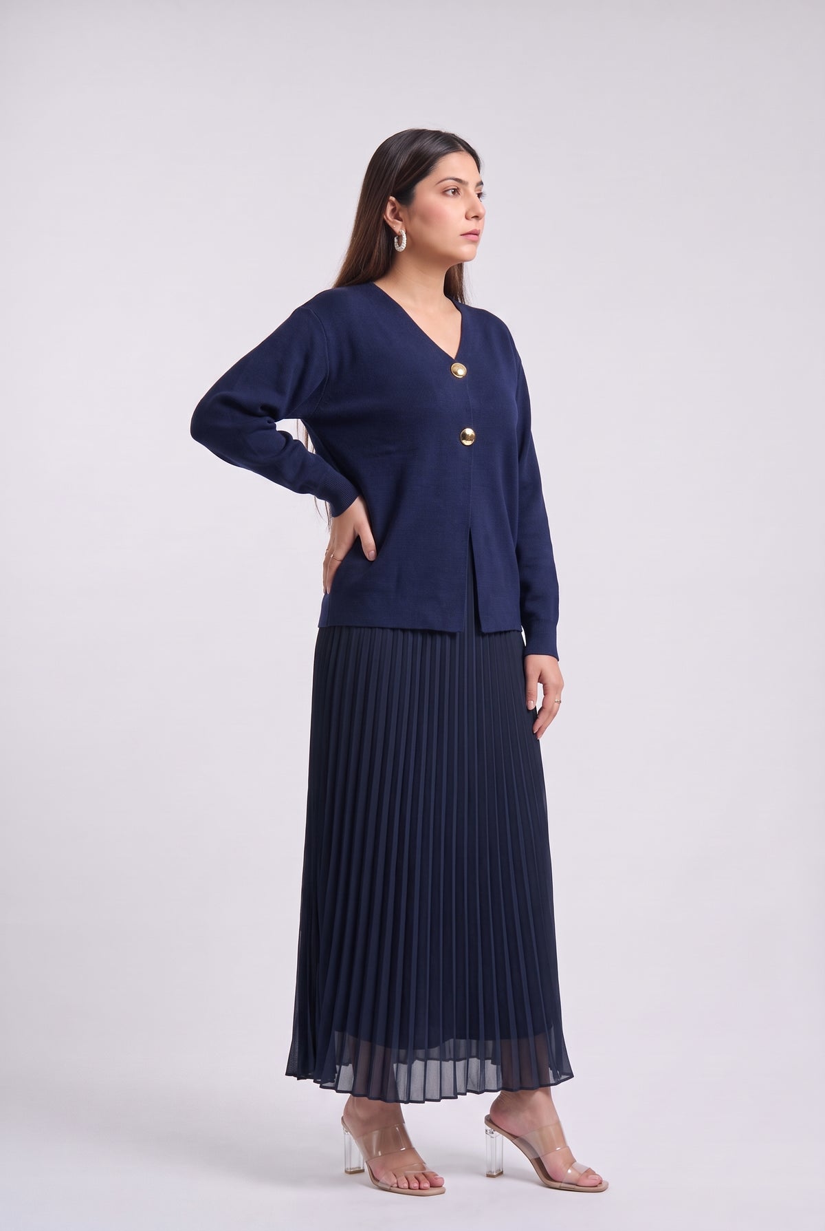 A woman modeling a navy blue two-piece outfit consisting of a long-sleeve button-front cardigan and a matching pleated maxi skirt, paired with clear high-heeled sandals against a light gray studio background.
