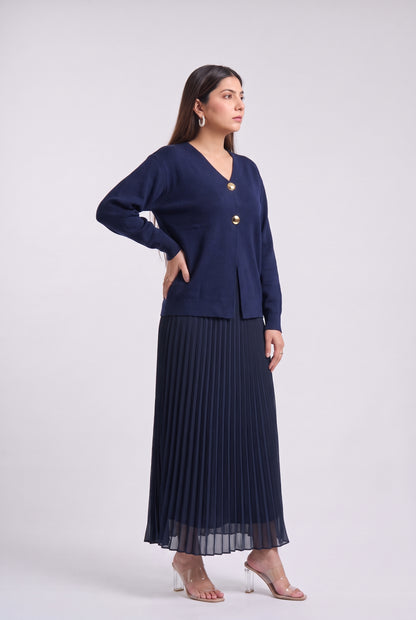 A woman modeling a navy blue two-piece outfit consisting of a long-sleeve button-front cardigan and a matching pleated maxi skirt, paired with clear high-heeled sandals against a light gray studio background.