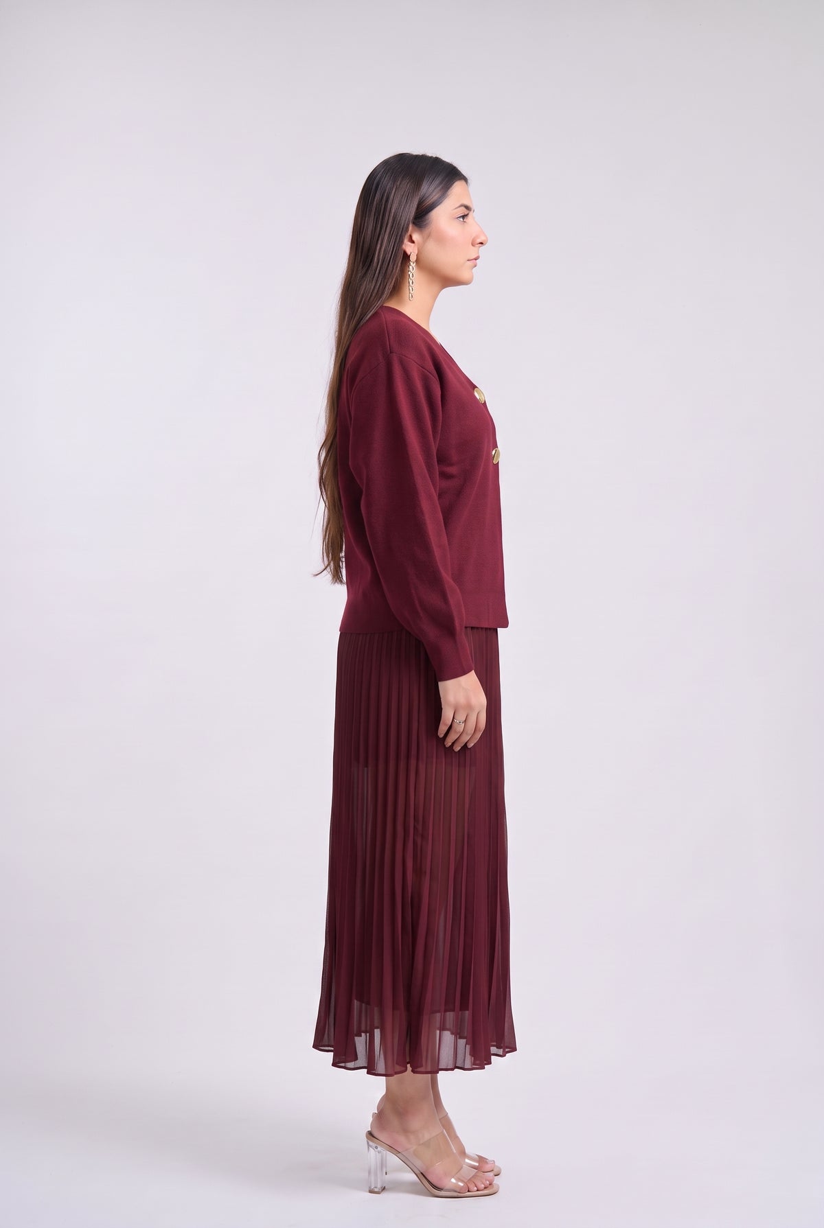 A woman in profile wearing a long-sleeve maroon knit top and matching pleated maxi skirt, paired with clear high-heeled sandals, against a light gray studio background.