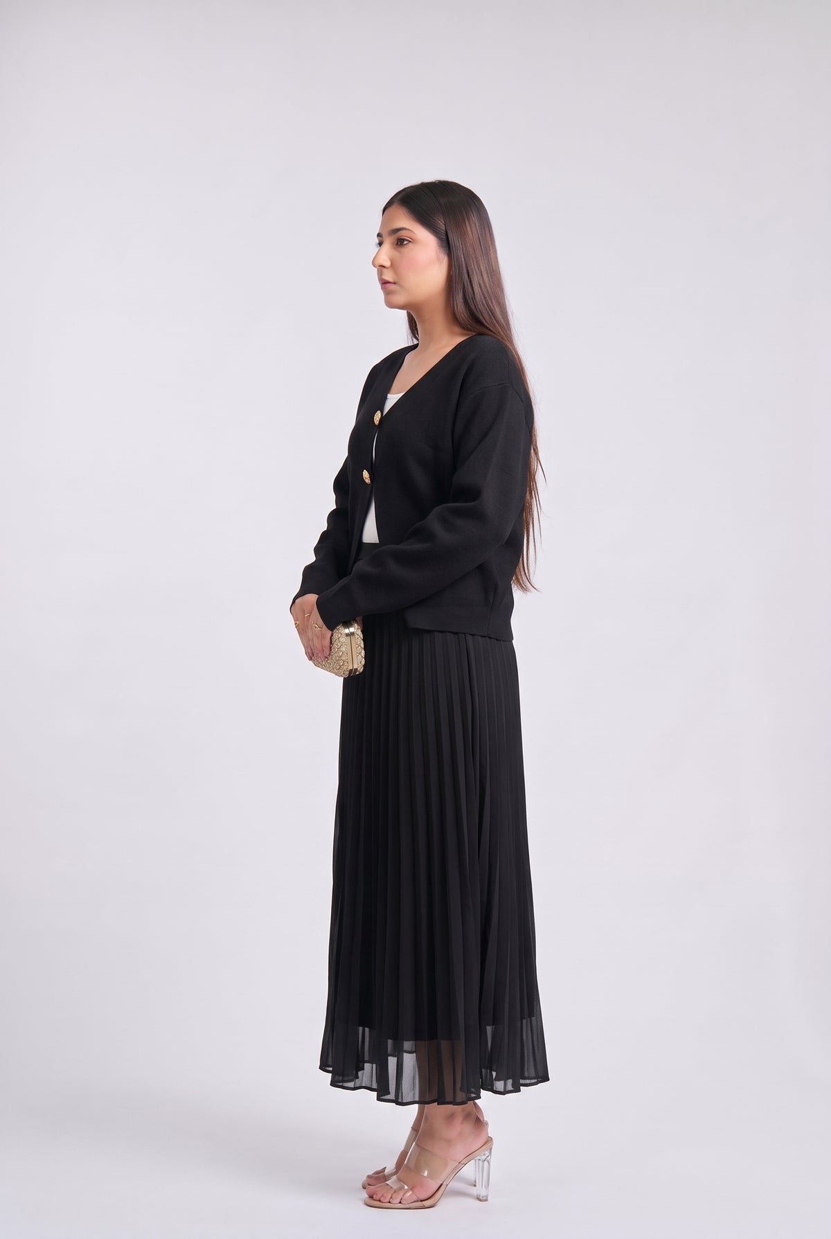 A woman stands in profile wearing a black long-sleeve knit cardigan over a white top and a long black pleated skirt, holding a small gold clutch against a plain light gray studio background.
