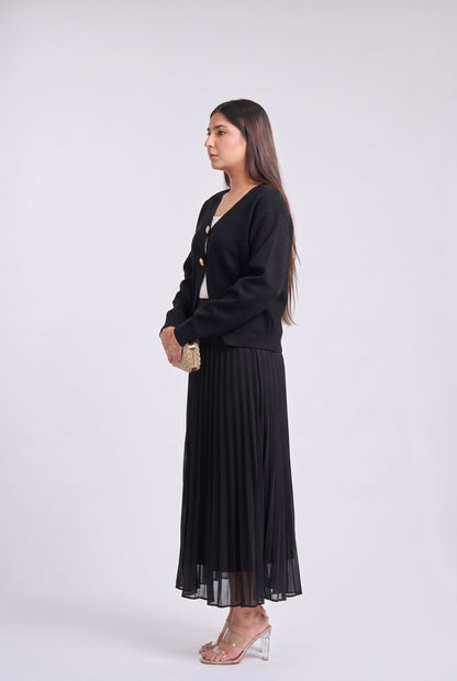 A woman stands in profile wearing a black long-sleeve knit cardigan over a white top and a long black pleated skirt, holding a small gold clutch against a plain light gray studio background.