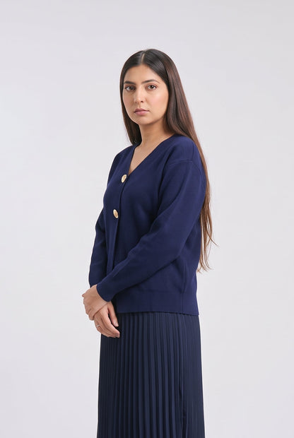 A woman modeling a navy blue cardigan with gold buttons and a matching navy pleated skirt against a light gray studio background.