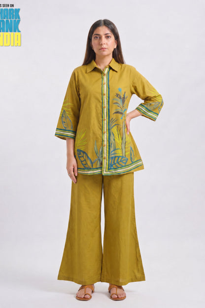 A woman modeling a mustard yellow two-piece kurta set with green and blue botanical embroidery, featuring a button-up shirt and matching wide-leg trousers, set against a plain light gray background.