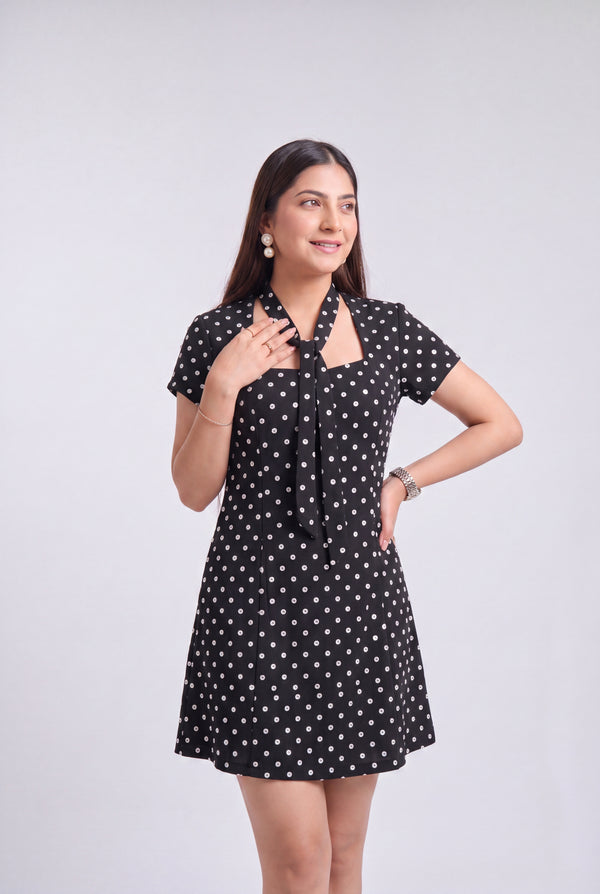 A woman modeling a short-sleeve black dress with a white polka dot pattern, featuring a tie-neck detail and a flared skirt, set against a plain light gray background.