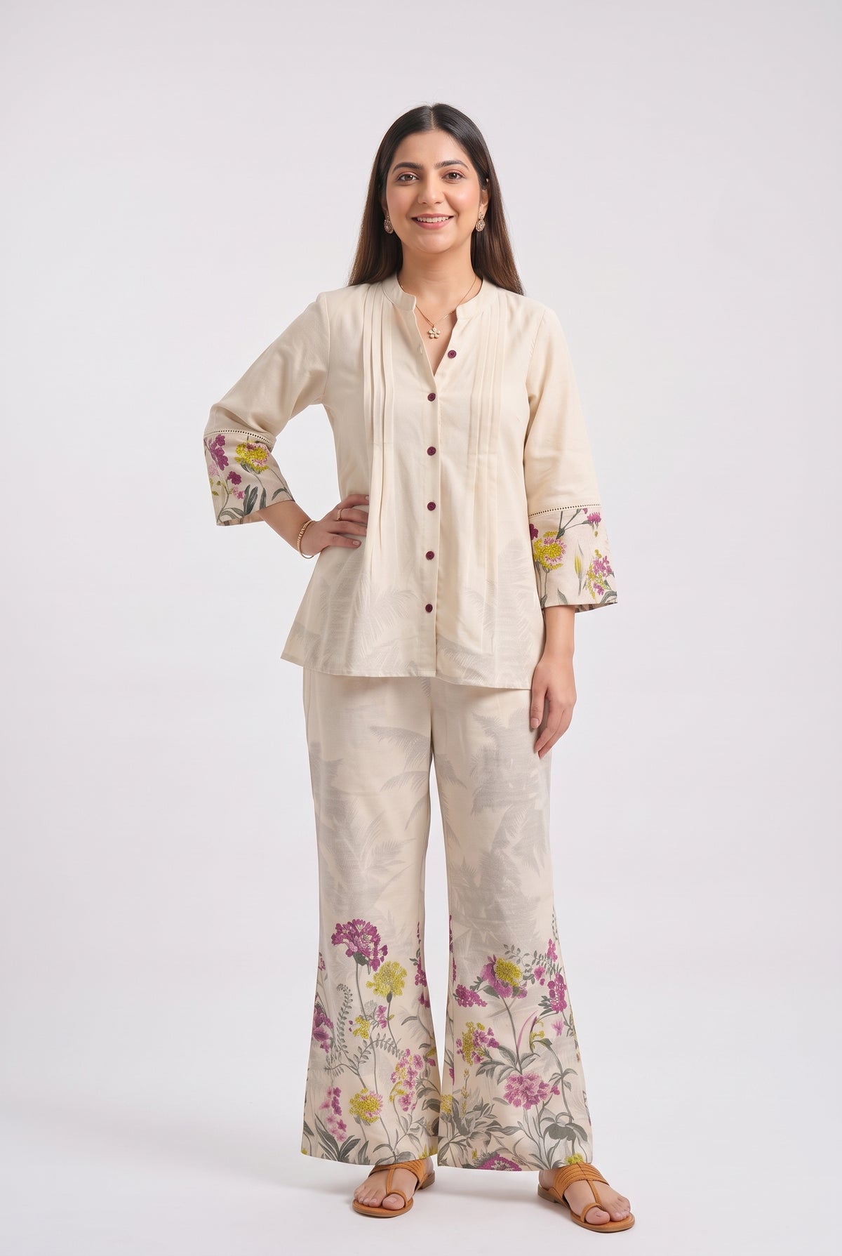 A woman modeling a two-piece cream-colored linen pajama set with floral prints on the cuffs and pant hems, photographed against a solid light gray background.